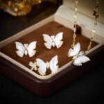 Wholesale Butterfly Shell Jewelry Set shown in gold and silver colors. The elegant set includes a butterfly pendant necklace, stud earrings, and an adjustable ring, all featuring iridescent Mother of Pearl wings and micro-paved Cubic Zirconia accents.