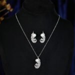 Wholesale Feather Pearl Zircon Set in a platinum-plated finish on a black display bust. Features a matching necklace and stud earrings, both in a curved feather shape paved with cubic zirconia, centered with a single white pearl.