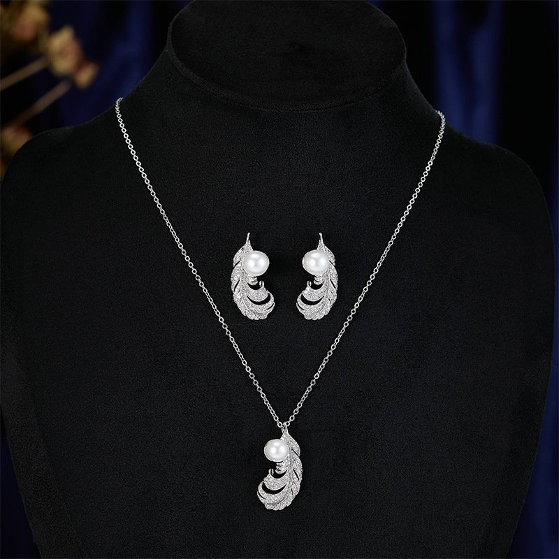 Wholesale Feather Pearl Zircon Set in a platinum-plated finish on a black display bust. Features a matching necklace and stud earrings, both in a curved feather shape paved with cubic zirconia, centered with a single white pearl.
