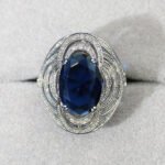 Wholesale blue zircon ring with oval-cut center stone and tiered halo design on gold plated copper from JuneJewelry.com – adjustable cocktail jewelry for bulk orders.