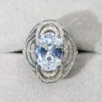 Wholesale blue zircon ring with oval-cut center stone and tiered halo design on gold plated copper from JuneJewelry.com – adjustable cocktail jewelry for bulk orders.
