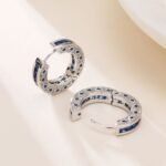 Sapphire blue huggie hoop earrings featuring channel-set princess-cut blue cubic zirconia, side filigree lattice detailing, and platinum plating.