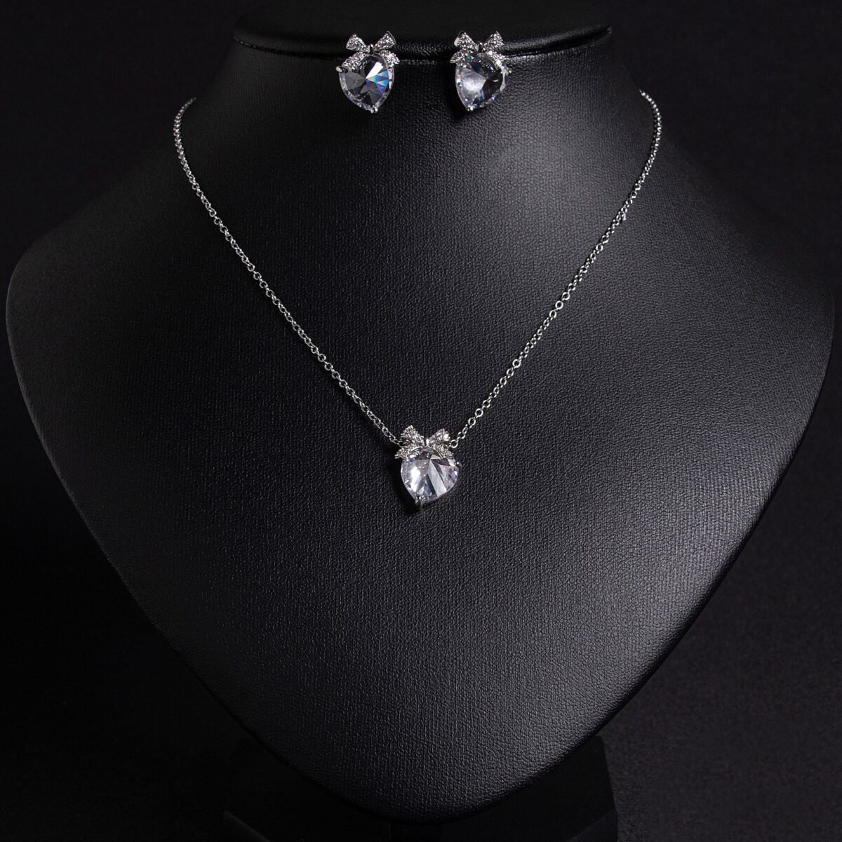 Niche bow heart crystal jewelry set wholesale featuring a delicate pave CZ bow ribbon knot topped heart crystal pendant chain necklace and matching stud earrings from junejewelry.com
