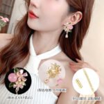 Ornate botanical dangle earrings featuring dual crystal flower blooms, glossy green enamel leaves, a smooth round bead, and a dangling teardrop CZ in silver and gold metal finishes.