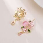 Ornate botanical dangle earrings featuring dual crystal flower blooms, glossy green enamel leaves, a smooth round bead, and a dangling teardrop CZ in silver and gold metal finishes.