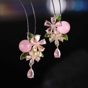 Ornate botanical dangle earrings featuring dual crystal flower blooms, glossy green enamel leaves, a smooth round bead, and a dangling teardrop CZ in silver and gold metal finishes.
