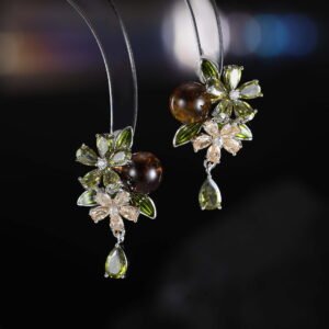 Ornate botanical dangle earrings featuring dual crystal flower blooms, glossy green enamel leaves, a smooth round bead, and a dangling teardrop CZ in silver and gold metal finishes.