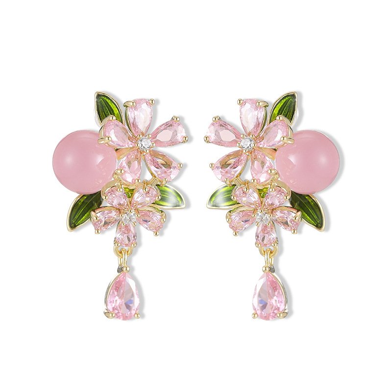 Ornate botanical dangle earrings featuring dual crystal flower blooms, glossy green enamel leaves, a smooth round bead, and a dangling teardrop CZ in silver and gold metal finishes.