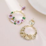 Twisted round hoop earrings adorned with hand-painted enamel flowers, green leaves, and CZ crystal accents in purple and white options.