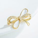 Delicate bow knot shell brooch wholesale featuring pavé cubic zirconia ribbons, inlaid natural mother of pearl shell loops, and a gold-plated finish from junejewelry.com