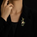 Playful Chinese style hanging panda bamboo CZ brooch wholesale featuring a chubby micro-pave cubic zirconia panda clinging to a gold-plated bamboo stalk with vibrant green enamel leaves from junejewelry.com