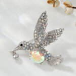 Hummingbird crystal brooch wholesale featuring an iridescent oval AB glass stone center, rainbow rhinestone-paved wings, and a dangling crystal beak charm in silver plating from junejewelry.com