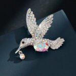 Hummingbird crystal brooch wholesale featuring an iridescent oval AB glass stone center, rainbow rhinestone-paved wings, and a dangling crystal beak charm in silver plating from junejewelry.com