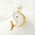 Playful cute mother of pearl whale brooch wholesale featuring a smooth iridescent white shell body, black gem eye, and round pearl water spout in gold or silver trim from junejewelry.com