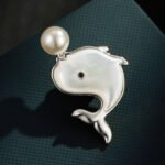 Playful cute mother of pearl whale brooch wholesale featuring a smooth iridescent white shell body, black gem eye, and round pearl water spout in gold or silver trim from junejewelry.com