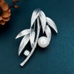 Elegant bamboo leaf pearl brooch wholesale featuring textured metallic leaves, a pavé cubic zirconia curved bamboo stalk, a sparkling solitaire round CZ accent, and a glossy pearl drop from junejewelry.com