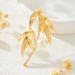 Elegant bamboo leaf pearl brooch wholesale featuring textured metallic leaves, a pavé cubic zirconia curved bamboo stalk, a sparkling solitaire round CZ accent, and a glossy pearl drop from junejewelry.com