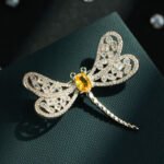 Dragonfly brooch wholesale featuring openwork filigree wings set with clear cubic zirconia, an oval gemstone body, and green CZ eyes from junejewelry.com