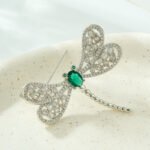 Dragonfly brooch wholesale featuring openwork filigree wings set with clear cubic zirconia, an oval gemstone body, and green CZ eyes from junejewelry.com