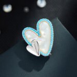 Double heart mother of pearl brooch wholesale featuring a carved shell primary heart with a turquoise or clear CZ halo border alongside a metallic accent heart from junejewelry.com