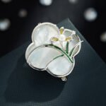 Vintage style four leaf clover mother of pearl orchid brooch wholesale featuring a natural white mother of pearl clover base overlaid with green enamel orchid leaves and a freshwater pearl flower center in gold or silver finish from junejewelry.com