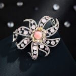 Light luxury pink floral queen conch shell brooch wholesale featuring a pink queen conch shell central cabochon surrounded by yellow, pink, clear cubic zirconia stones, soft purple bead accents, and silver pave openwork petals from junejewelry.com