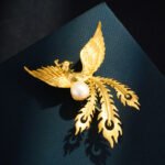 Chinese style brushed golden phoenix pearl brooch wholesale featuring wire-brushed gold feathers, outstretched wings, flowing tail plumes, and a central white freshwater pearl from junejewelry.com.